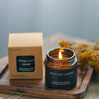 Scented candle with a lit flame next to its packaging on a wooden surface.